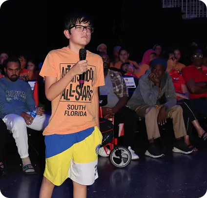 Boy with orange shirt holding a microphone in front of crowd